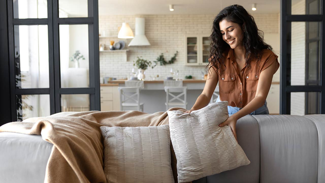 Lovely young woman putting soft pillows and plaid on comfy sofa, making her home cozy and warm