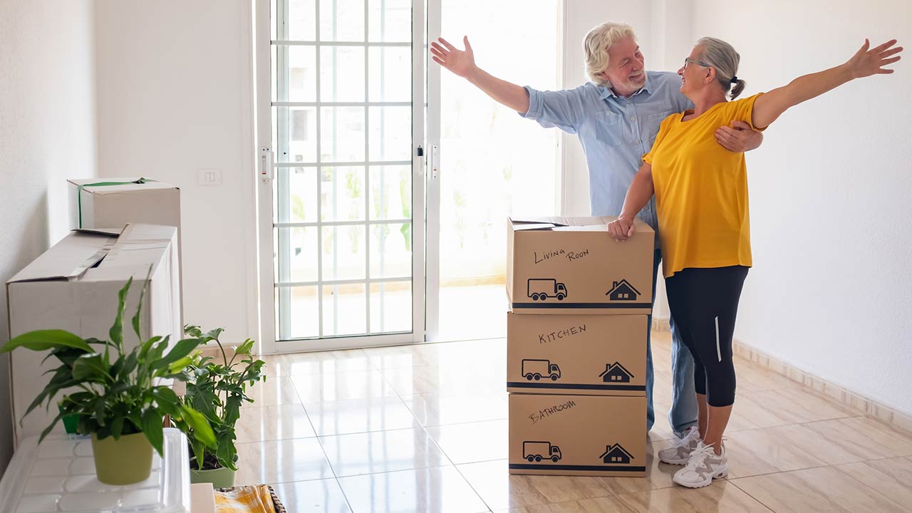 Smiling caucasian senior couple white haired open arms looking at each other's eyes during relocation, excited for a new beginning