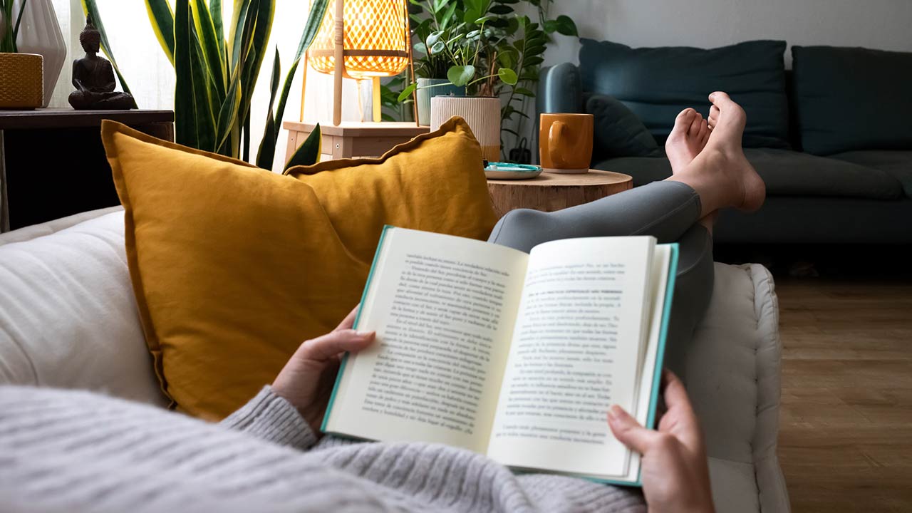 young woman relaxing at home reading a book lying on sofa