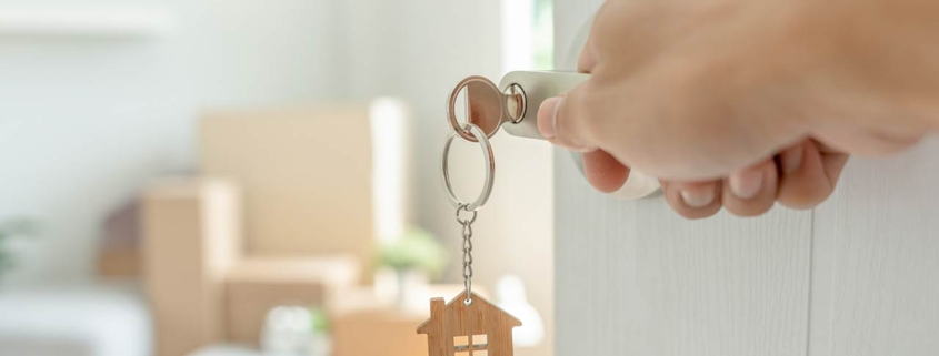 A key inserted into a door of the new house with a hand opening it, inside the room are cardboard boxes and furniture