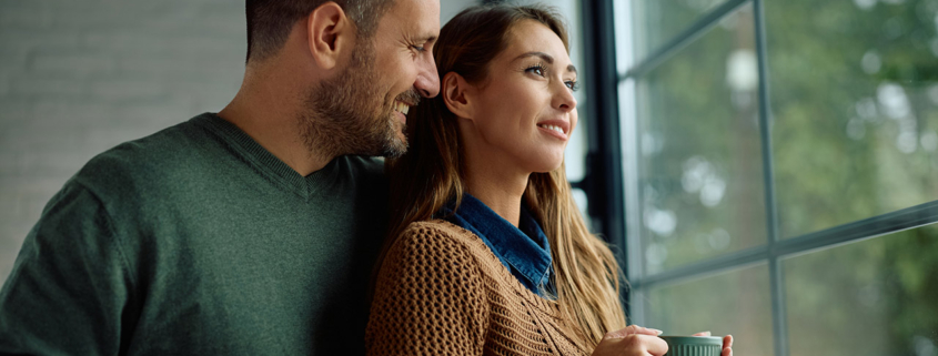 Smiling woman day dreaming while drinking coffee with her husband by the window at home